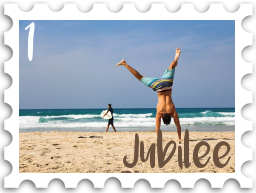 January 2025 Jubilee SWG challenge stamp - photo of a man in long swim trunks doing a cartwheel on the beach on a sunny day. A surfer is entering the water in the background. 
