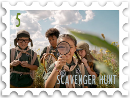 May 2026 Scavenger Hunt SWG challenge  stamp - a color photo of a group of children in what may be scouting uniforms out in a green field on a sunny day. The child in the center holds a large magnifying glass which she peers through at the viewer. The other children are clustered behind and around her, looking intent.