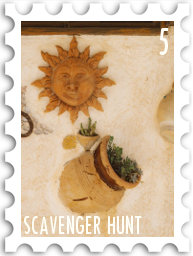 May 2026 Scavenger Hunt SWG challenge  stamp - a color photo of a stylized sculpture of the sun hanging on a stucco wall above several embedded planters from which greenery spills. A yellow shell is also embedded in the wall.  The scene is warm and cheerful.