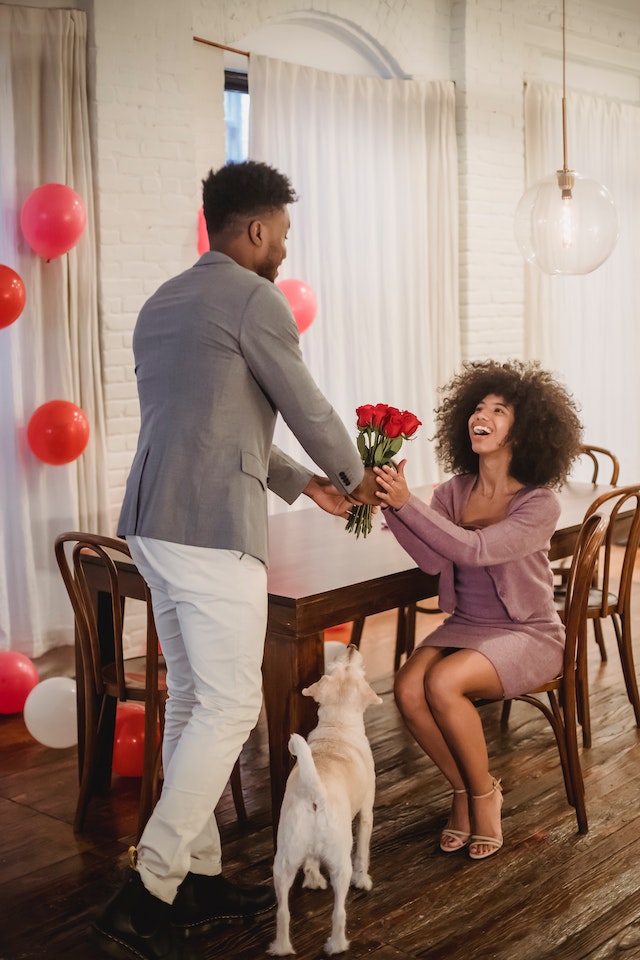 A Black man hands red roses to a smiling Black woman seated at a table; the room is decorated with red and pink balloons and a dog gazes up at the couple