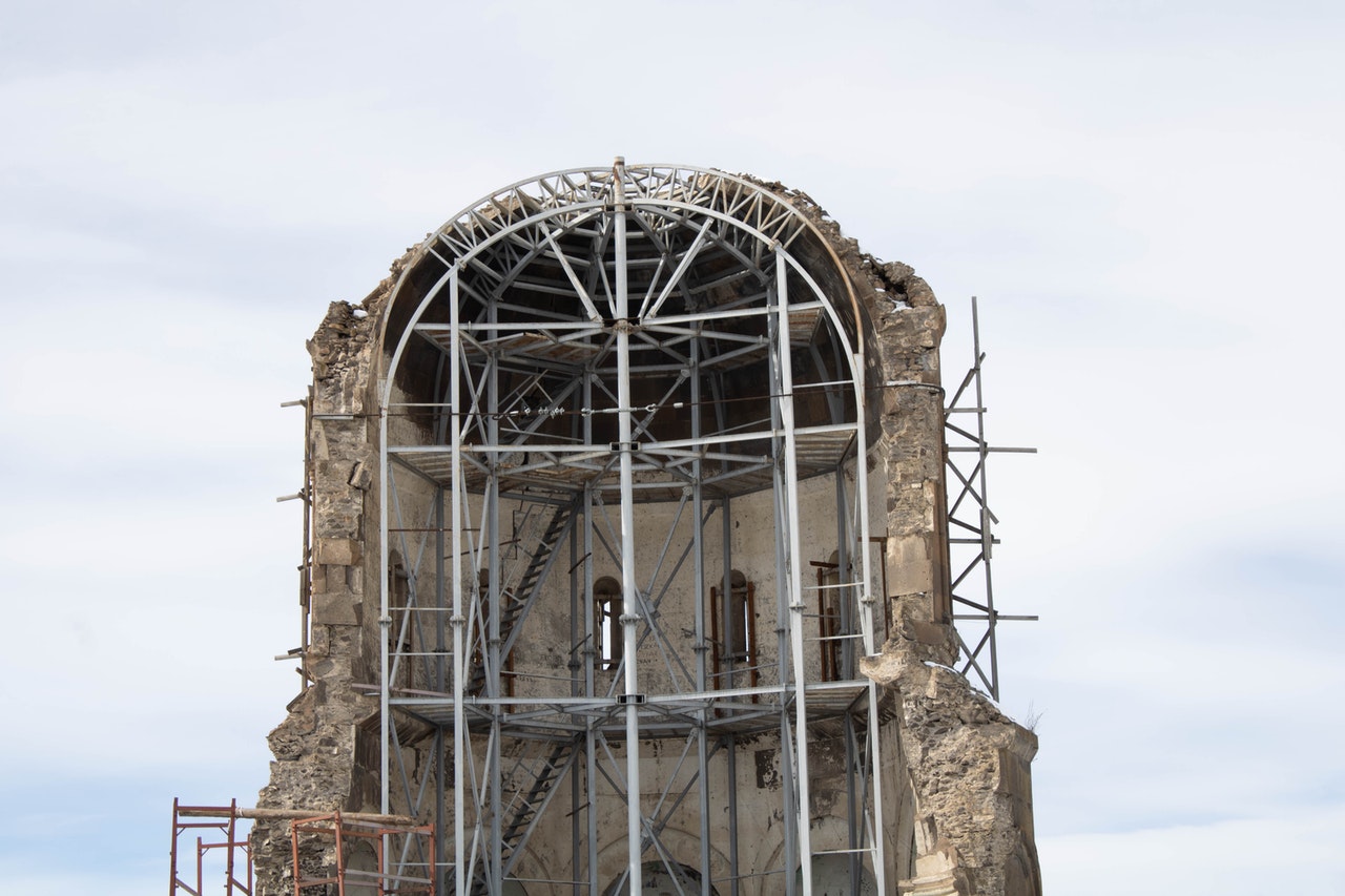 The outer shell of a domed building, possibly a tower, supported from both sides by a skeleton of metal scaffolding. Half the building has crumbled or collapsed, leaving an almost perfect cutaway view.