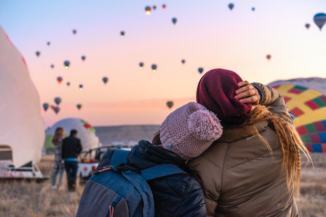 Two women sit close and watch hot air balloons launch; one woman is resting her head on the other's shoulder