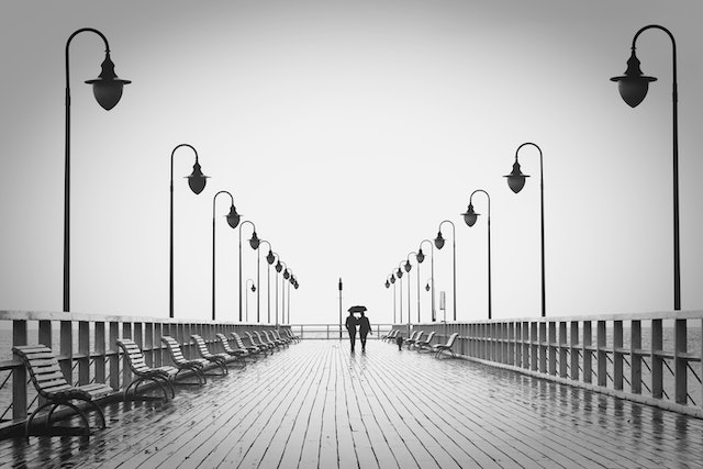 A black-and-white photo of a couple in the distance, walking on a pier toward the sea