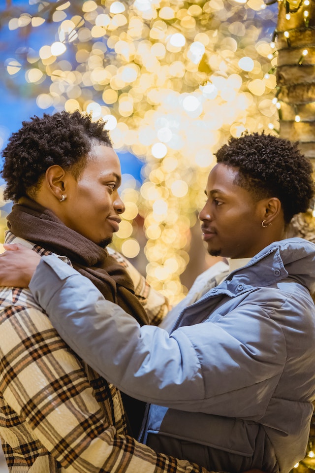 A Black man and a Black woman embrace and gaze into each others' eyes with sparkling lights in the background