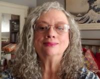 Woman in her 60s with long, silver curly hair and glasses