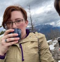 A person of indiscriminate gender sits on top of a mountain. They wear a tan jacket and are holding up a blue teacup in front of their face. They have short, side-swept red hair and rectangular glasses.