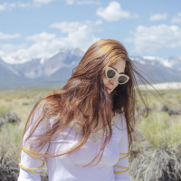 woman with golden-brown hair standing in front of some mountains wearing sunglasses