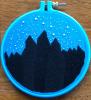 a piece of embroidery on blue fabric in a blue hoop, featuring the black silhouettes of mountains with three taller peaks in the middle, and above them stars in french knots, with larger knots depicting the valacirca/big dipper