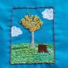 an embroidery on blue fabric of a dark brown tree stump and a lighter colored young tree on a green hill dotted with blue and yellow flowers; the tree has many yellow flowers and a few dark green leaves; there are two clouds in the sky background, and it is enclosed in a square border of dusty pink 