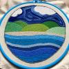an embroidered scene of water in many shades of blue, a pale tan strech of beach, and five green hills under a variagated blue sky and two white clouds, all in chain stitch on an off-white background fabric