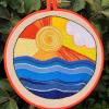 a round embroidery hoop on a background of green leaves; the embroidered scene is a sunset in yellows, reds, and oranges with a white cloud, and varying shades of blue on the bottom half for the sea, all done in chain stitch; the scene is bordered with gray backstitch