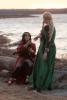 Photo of Maglor and Galadriel cosplayers. Maglor is sitting on a rock by the water's edge, eyes wide open, reaching a hand out to Galadriel, who stands with her back to the camera, reaching out to Maglor's hand but not touching it. The sun is setting and the colors are faded. Maglor wears a red robe, red capelet, brown tabard, gray tunic, black pants and black and red embroidered shoes. He has silver eyes, thick black curly hair and a gold leaf circlet with a red teardrop jewel in the center. His hair blows