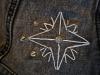 An eight pointed star embroidered on a grey jean jacket in white thread with three metal studs at the top/left side