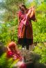 Cosplay photo taken in the woods. Maglor stands on a rock, gazing downward at Maedhros while singing and playing the lyre. Maedhros sits at the foot of the rock looking up at Maglor. Maedhros wears a red jacquard robe with silver leaf-patterned accents and silver-colored sleeves, and has a red and silver circlet. Maglor wears a long red robe and capelet with gold and black embroidered swirls, and a gold laurel.