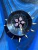 A glass flower floats surrounded by bubbles in an orb. Around the orb, at the points of the clock are 12 small wentletrap shells approximately 2-3cm long. The background is sparkly, swirly blues. Bright sunlight reflects off all the glass surfaces and refracts within the orb.  