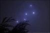 The constellation sometimes known as Cassiopeia in the night sky, with a tree branch in the bottom corner that appears to be cedar.  The air is very misty, making the stars glow with a faint bluish-purple haze. 